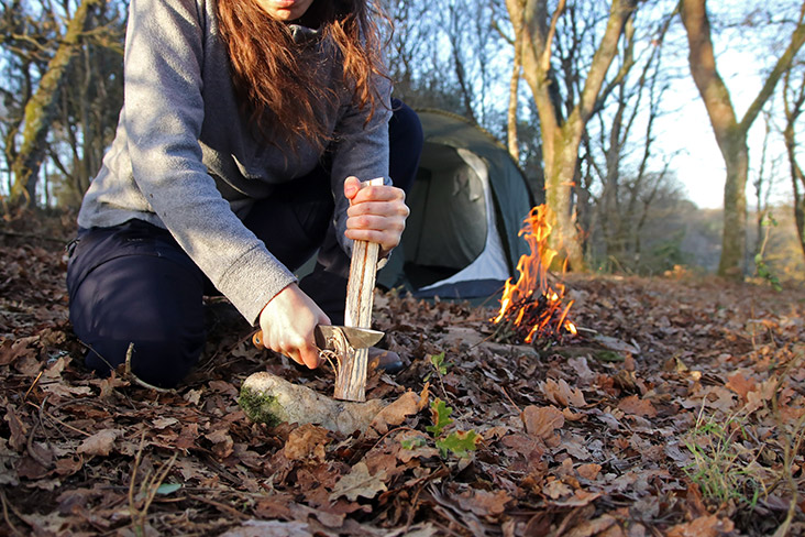 A woman is kneeling on a pile of autumn leaves and is cutting a piece of wood with a knife to make a feather stick. 
