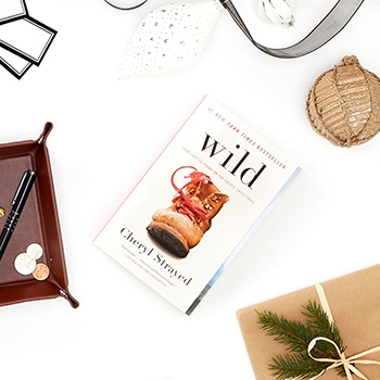 The book Wild by Cheryl Strayed, as viewed from above. On the book’s cover is a brown hiking boot with red shoe laces. An ornament and holiday ribbon placed near the book provide a festive look.