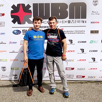 Panagiotis and his father, Ioannis, standing side by side and smiling in front of a backdrop with a WBM logo at Wicked Big Meet in 2018.