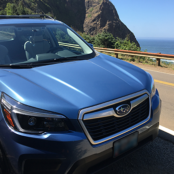 A front passenger side view of Buckle’s Subaru Forester parked along a roadway with the ocean shoreline and rocky cliffs behind the vehicle.