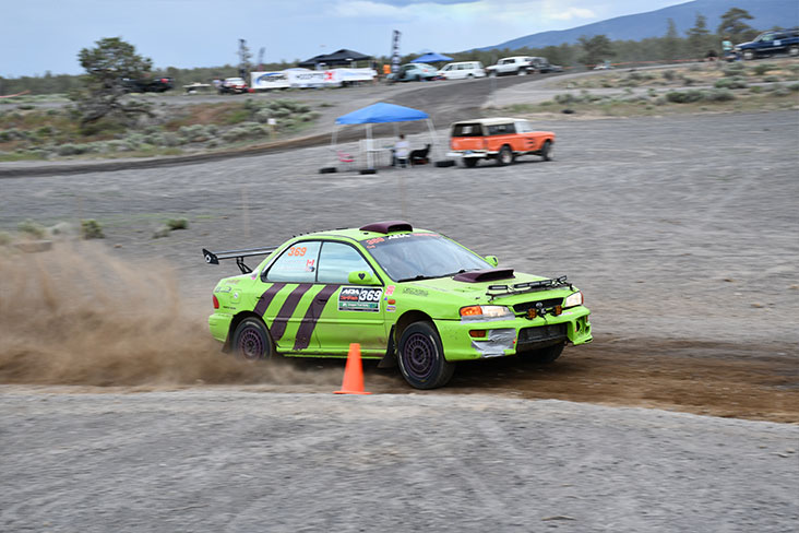 A light-green Subaru WRX races past an orange cone on a dirt road. 