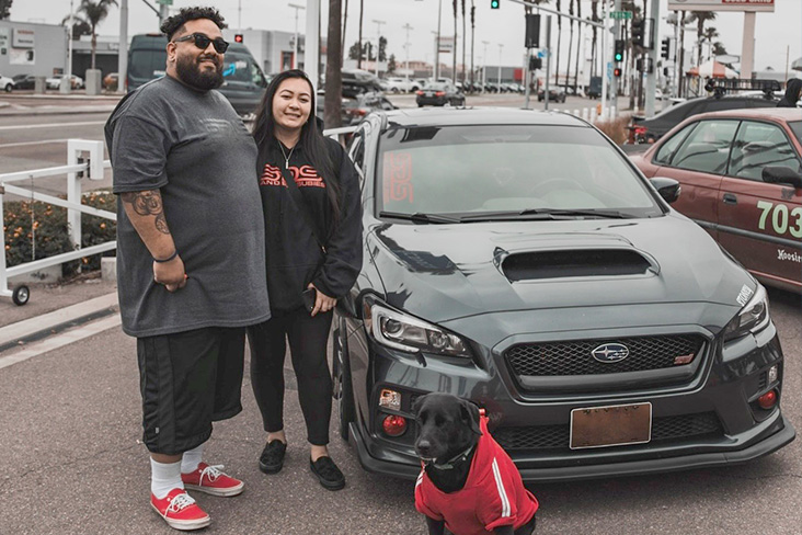 Rosie Le is standing with her husband, Michael Reyes, next to Rosie's 2017 Subaru WRX STI in a lot overlooking a busy city street.
