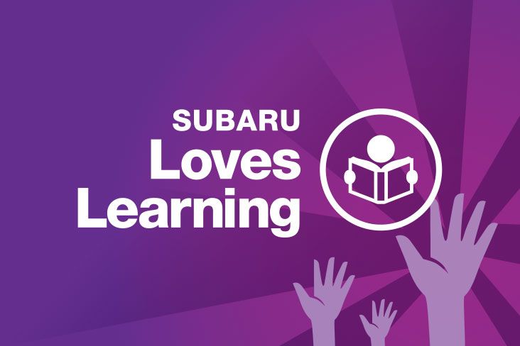 Subaru Loves Learning logo in white lettering on a purple background. To the right of the words “Subaru Loves Learning” is an illustration of a person reading a book, which is enclosed in a circle. Below it, hands are shown reaching up toward the circle.