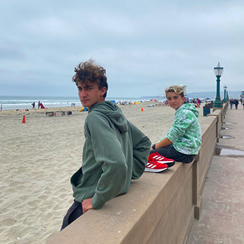 15-year-old Kai stands against a cement wall dividing the walkway from the beach. To Kai's left, his 11-year-old brother, Nikko, is seated on the wall. The sky is overcast and the beach is largely deserted.