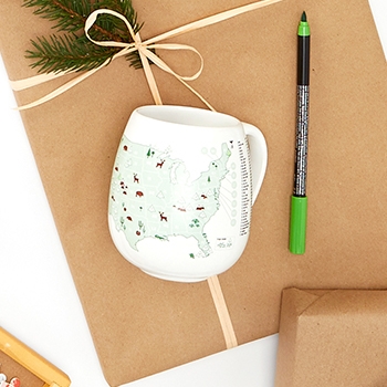 A white ceramic mug with a map of the United States on the side of it. Various locations on the map have been colored in using assorted colors. Next to the mug is a green ceramic marker. Decorative pine cones and pine needles placed near the mug provide a festive look.
