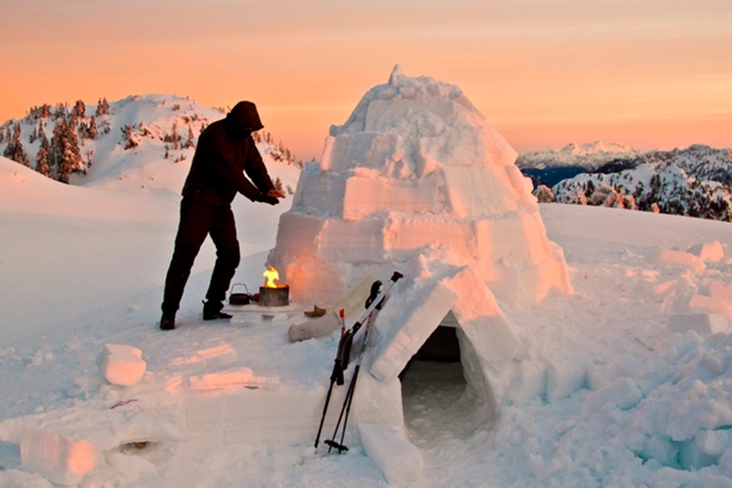 Snowy scene with male camper warming hands over a small fire next to a quinzee.