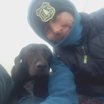 A closeup of Katy Nelson, who is wearing a U.S. Forest Service knit hat and a jacket, with her dog, Sadie.