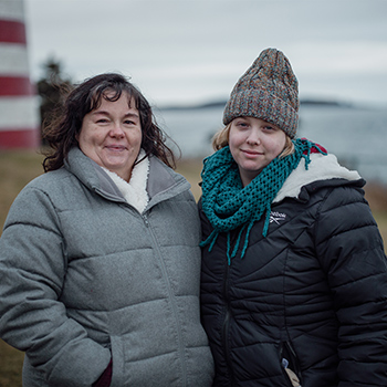Terrie McConnell is on the left, and Jamie McConnell is on the right. They are bundled up in winter gear, and the Maine coast is blurred in the background. 
