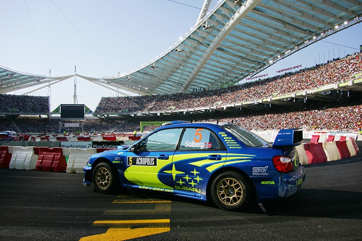 Petter Solberg driving a Subaru rally car at the Acropolis Rally. There is stadium seating to the right with hundreds of people in the stands. 