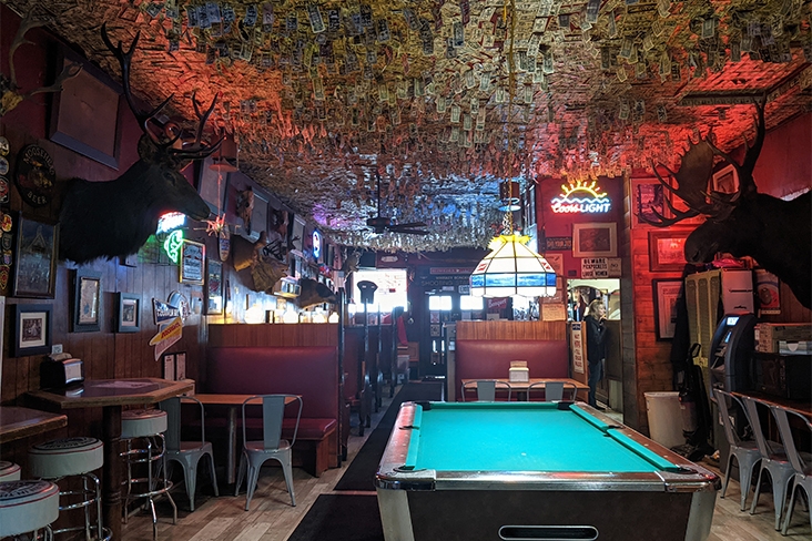 An interior shot of the Shooting Star Saloon in Huntsville, Utah. The room is low lit. There is a pool table in the center of the room, and on the wall, a moose head and deer bust with antlers are hung. There are several bar stools under small tables around the room and a few booths.