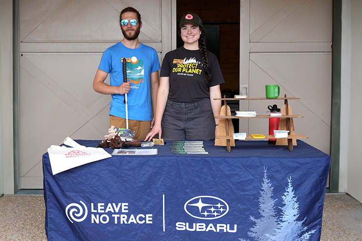 The McGraws are standing behind a Subaru and Leave No Trace table. Luke is on the left and is holding a white cane against his chest. Becca is on his right wearing a Protect Our Planet T-shirt in black with yellow lettering.