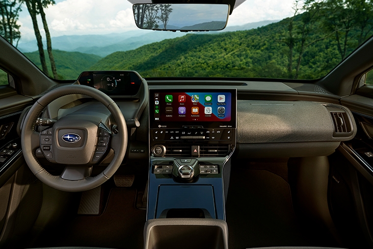 A full view of the Subaru Solterra cabin, with a large 12.3-inch touchscreen in the center and views through the windshield of green tree-covered mountains.