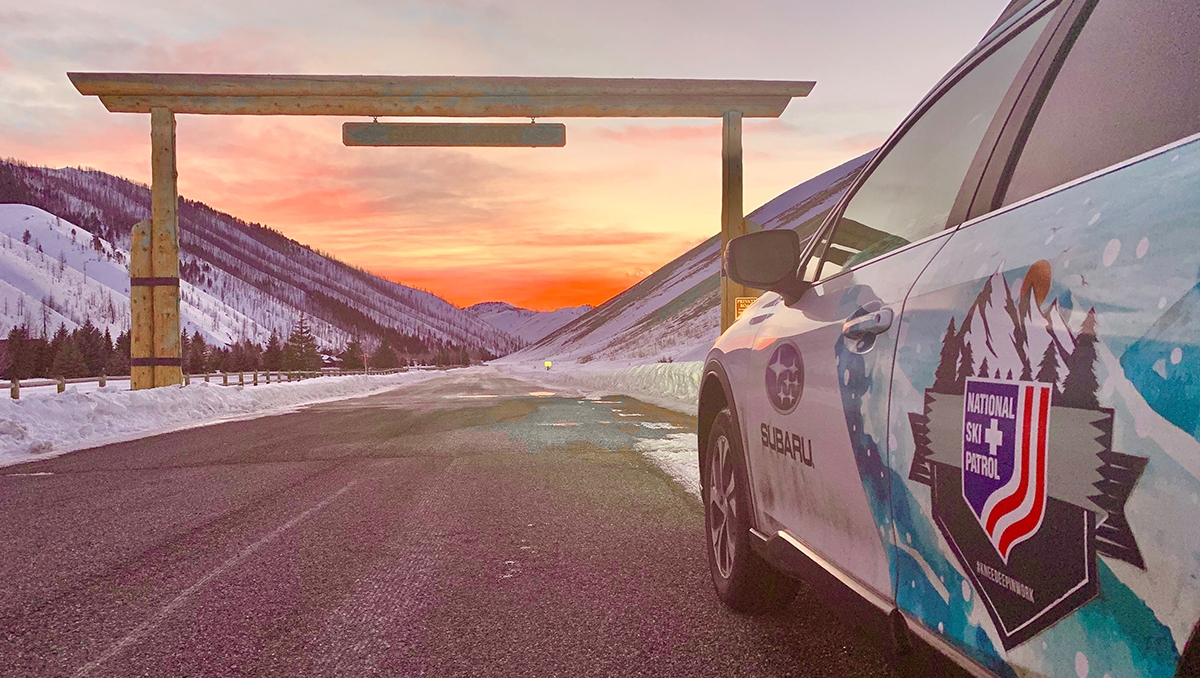 A Subaru Forester bearing the National Ski Patrol logo, parked in front of the entry to a ski area with an orange sunset in the background