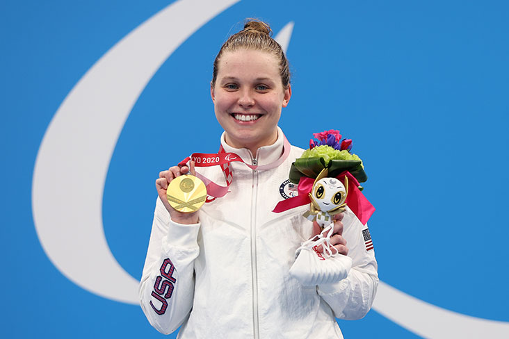 Morgan Stickney holding a gold medal at the 2020 Paralympic Games in Tokyo, Japan. She is wearing a white jacket that says USA on the sleeve and is smiling.