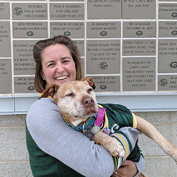Stacy Dubuc is carrying Ginger in her arms in front of a memorial wall at Lambeau Field in Green Bay, Wisconsin. Stacy is smiling, and Ginger looks relaxed and comfortable with her eyes partly closed.