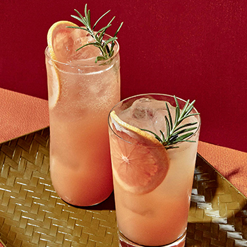 Two tall glasses are filled with a pink-colored beverage, grapefruit slices and ice. Each drink is topped off with a sprig of rosemary.  