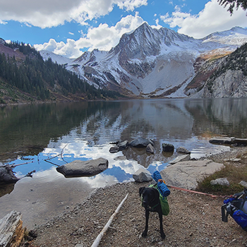 Sadie is standing near water with snow-capped mountains in the background. She is carrying a two-sided, lightweight backpack on her back. 