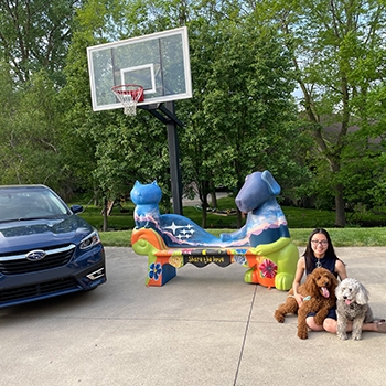 Madeline sitting with her two curly-haired dogs on her driveway with her painted loveseat in the background next to her Subaru Legacy Limited.