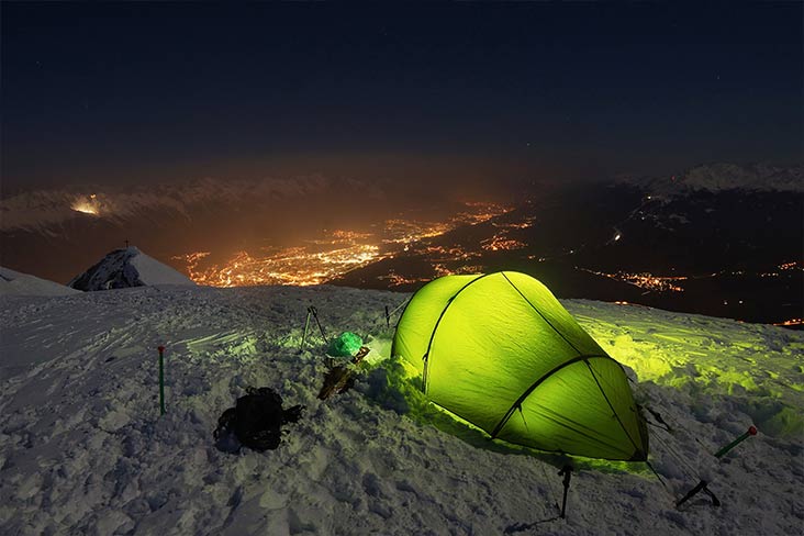 Nighttime winter camping scene with pitched tent on mountaintop in snow overlooking a city.