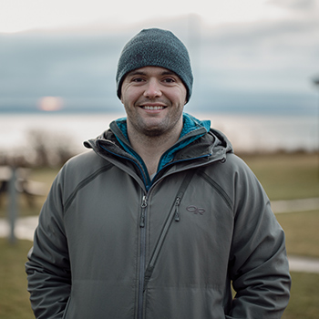A picture from the chest up of a smiling young man wearing a beanie and jacket with the Maine coast in the background