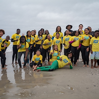 A large group of smiling adolescents and teens are standing or lying on the sand at the waterʼs edge. Theyʼre all wearing yellow T-shirts with the Laru Beya Collective logo.