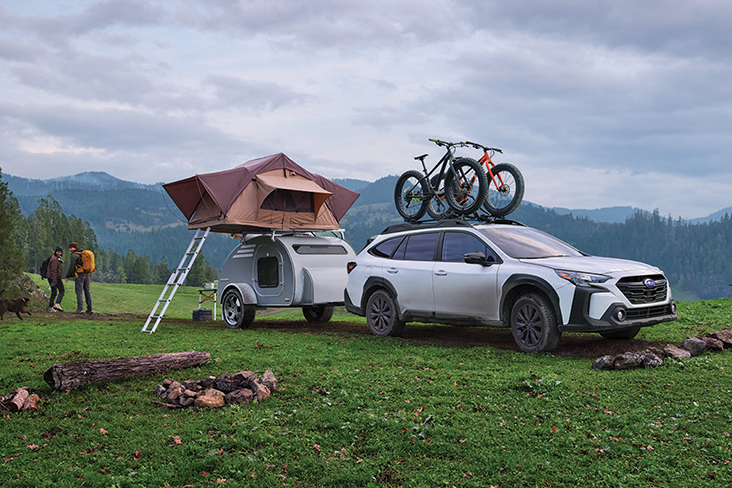 A 2025 Subaru Outback is parked in a grassy wilderness area with two bikes fastened in a bike rack on top. Behind the Outback is a small trailer, which has a fully extended ladder leading up to a rooftop tent that is assembled on top of the trailer. In the distance, two people with backpacks are hiking toward mountains.