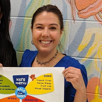Margaret Behrns is smiling and holding up a colorful lunch menu that says ‘kid’s menu’ and includes sides, desserts, and drinks. In the center of the menu is the Walk In My Shoes logo.