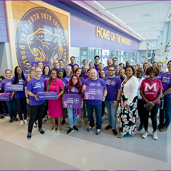 Subaru of America adopted all 55 classrooms at the four-school Camden High Campus in the automaker's hometown of Camden, New Jersey. Picture of about 30 people standing in an expansive school hallway, which says Home of the Panthers on the wall. Many in the group are wearing purple Subaru Loves Learning T-shirts. They are smiling and relaxed.