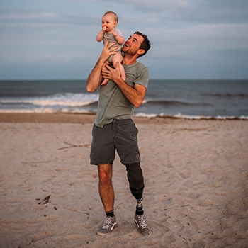 Joel, a white man, holds baby Land above his head on the beach. They are both smiling. Joel wears a grey T-shirt, dark grey shorts and sneakers. He has a prosthetic left leg.