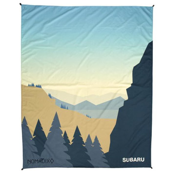 A Nomadix Festival Blanket with an illustrated forest scene in pastel colors. It says Nomadix on the bottom left corner, and on the bottom right corner, it says Subaru.