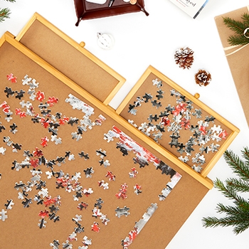 The Bits and Pieces wooden puzzle board seen from above; it has two drawers, which are open. A puzzle is started on the board and puzzle pieces are in one of the drawers. Decorative pine needles and pine cones placed near the board provide a festive look.