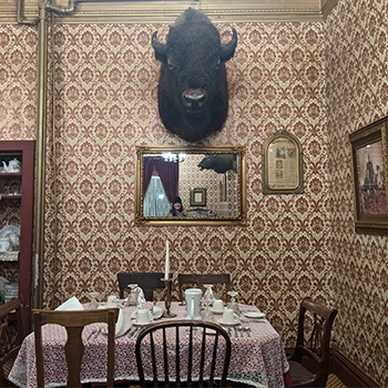 The dining room at The Historic Virginian Hotel has patterned wallpaper on all the walls. There is a large buffalo head mounted on the dominant wall, and below it is a rectangular dining table with white tableware on top of it and six mismatched wooden dining chairs set around the table.