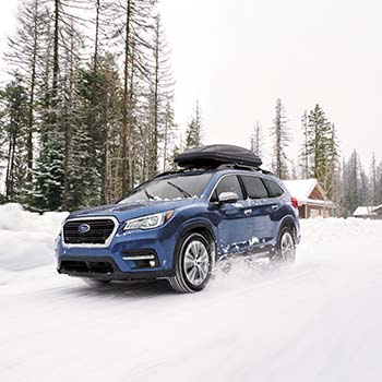 A 2022 Subaru Ascent in Abyss Blue Pearl is driving away from a cabin through several inches of snow with a Thule Cargo Carrier mounted securely on its rooftop.