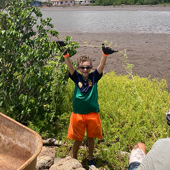 Nikko is holding up a long stem of pickleweed and smiling. Behind him is a muddy patch with houses in the distance.