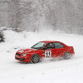 A red vehicle with the number 11 on the driver door is driving fast on a road. It’s snowing and leafless trees can be seen in the distance.