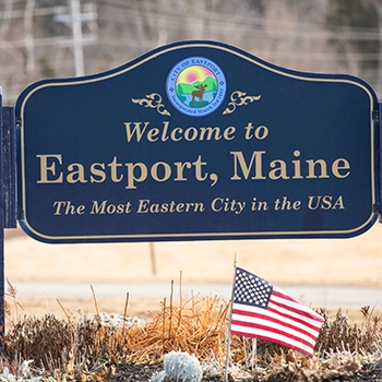 A closeup of a decorative navy-blue sign that reads, “Welcome to Eastport, Maine. The Most Eastern City in the USA.” A small American flag is stuck in the ground in front of the sign.