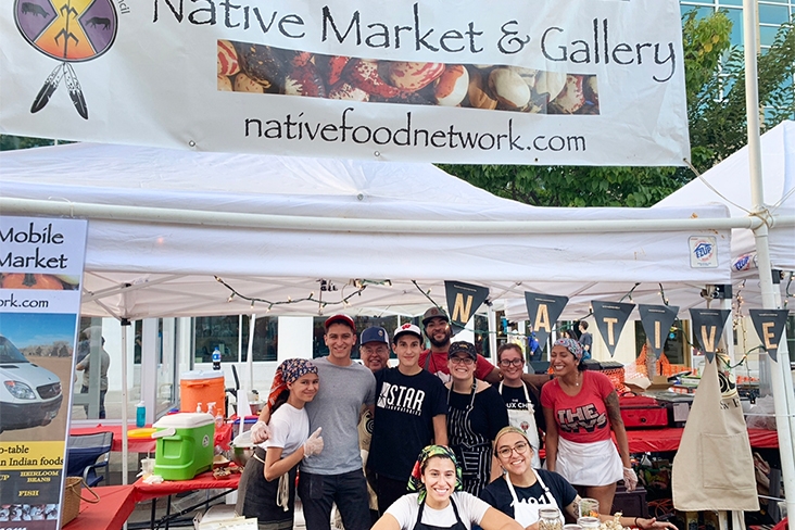 The Wild Bearies crew at the Taste of Madison in Madison, Wisconsin. Eight staff members, wearing headscarves or hats, are standing under a white tent, smiling. Above them is a sign that says, Native Market & Gallery, nativefoodnetwork.com. Two people are seated at a table in front of the tent. There are coolers on a table behind the group for food prep.