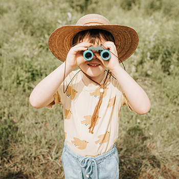 A child is outside ready for adventure wearing a straw hat and looking through a small set of plastic binoculars.