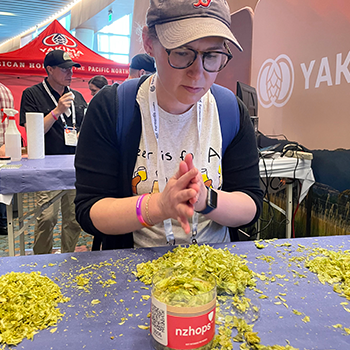 Sauter is standing behind a table with what appear to be hops spread out on the table. There is a container that says nzhops in the foreground on the table. She is concentrating. Other contestants are in the distance behind her.