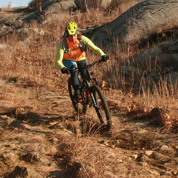 Subaru owner Mary Cryan is riding a bike on a rocky trail. She’s wearing a long-sleeved yellow shirt, neon orange vest, black athletic pants and a yellow protective helmet.