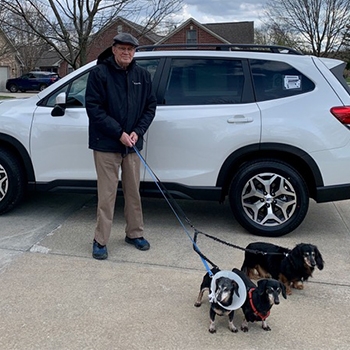 An older gentleman, wearing brown pants and a dark-colored jacket, is standing next to his Subaru Forester with the three dachshund boys on a leash.