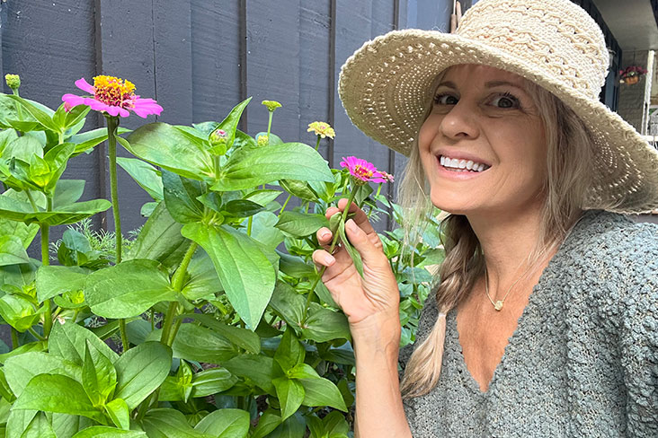 Writer Jennifer Fischer is standing next to her healthy-looking zinnia flowers, which are almost as tall as she is. Fischer is wearing a straw hat and is smiling as she holds a zinnia by the branch.