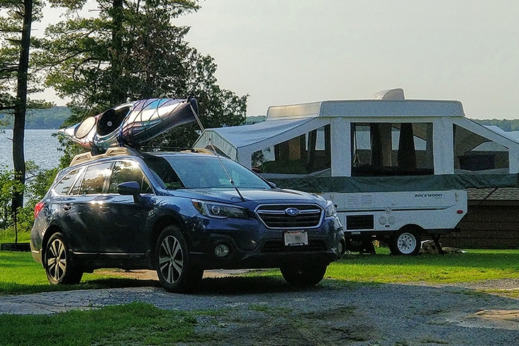 A 2019 Subaru Outback Limited with a kayak on the roof is parked at a campground with evergreens and lake water in the distance behind it. A pop-up camper is near the Outback.