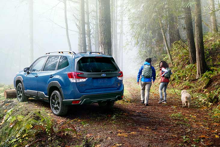 Two people are heading up a forest path with a small white dog on a leash, and a 2022 Subaru Forester is parked on the left. They are both wearing backpacks, and one person is turned toward the other, smiling.
