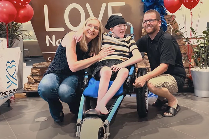 Colin Jolly is seated in his new off-road wheelchair. His parents, Kari and Dan, are kneeling beside him, smiling.