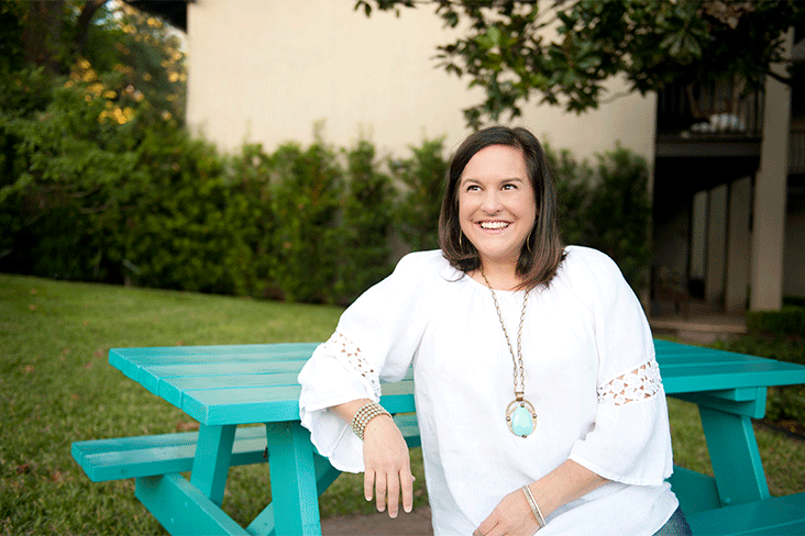 Kristin Schell, wearing a white blouse with three-quarter sleeves, is seated at her turquoise table, smiling.