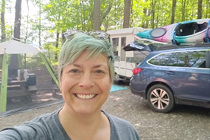 Kris Isaacson taking a selfie at her campsite in Taughannock Falls in New York state. Behind her on the left, a tent is set up with a picnic table inside it, and on the right is her Subaru Outback with a multi-colored kayak on top and a camper behind it.