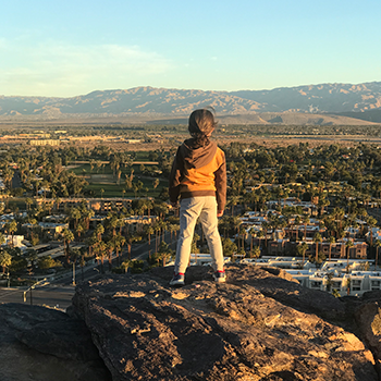 Everest Yasuda, seen from behind, as he stands on a large rock looking toward Palm Springs, California, from Rimrock Trail. Mountains can be seen in the far distance.