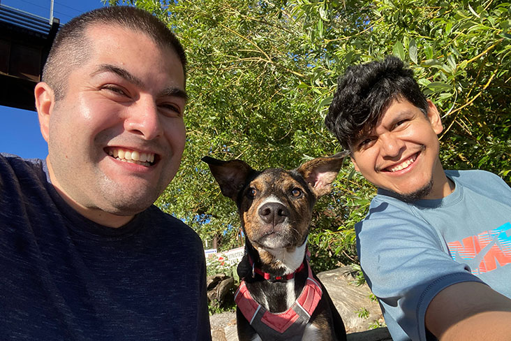Emmanuel Camarillo is smiling on the left-hand side, and Francisco Contreras Alvarez is smiling on the right. Between them is a medium-sized dog with large ears that is wearing a red collar and scarf. A deciduous tree and blue skies can be seen in the background. 