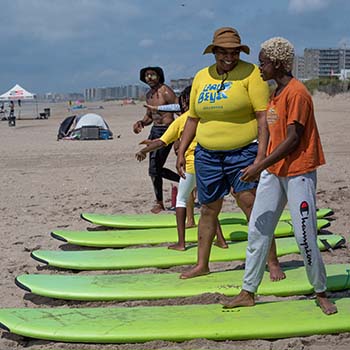 An instructor in a Laru Beya Collective T-shirt and wide-brimmed hat teaches a class. All the surfboards are lying flat on the sand, and there are three students in the cropped shot.